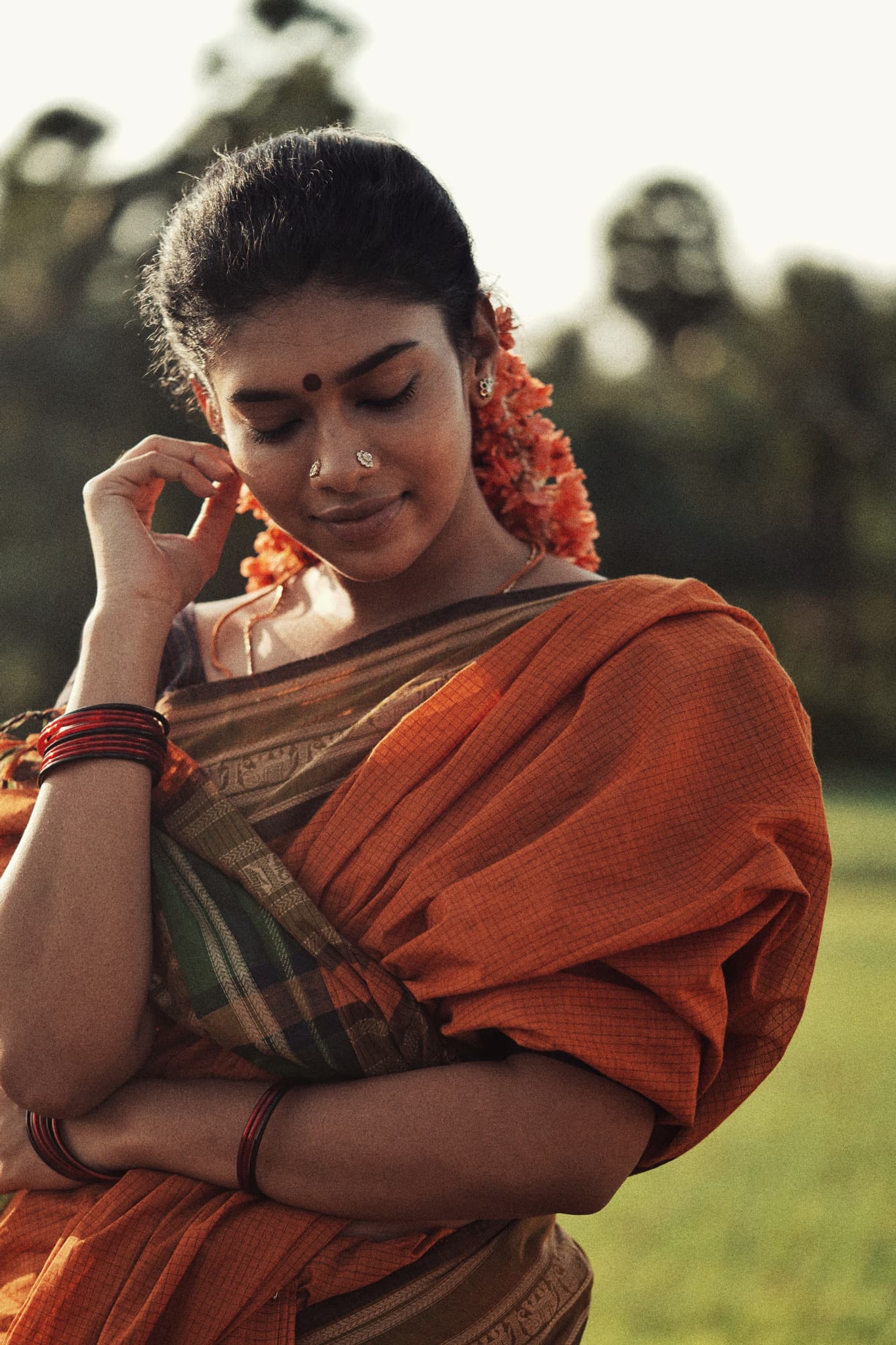 A woman wrapped in a rust-orange handloom saree, its woven border gathered at her arms, glass bangles at her wrists
